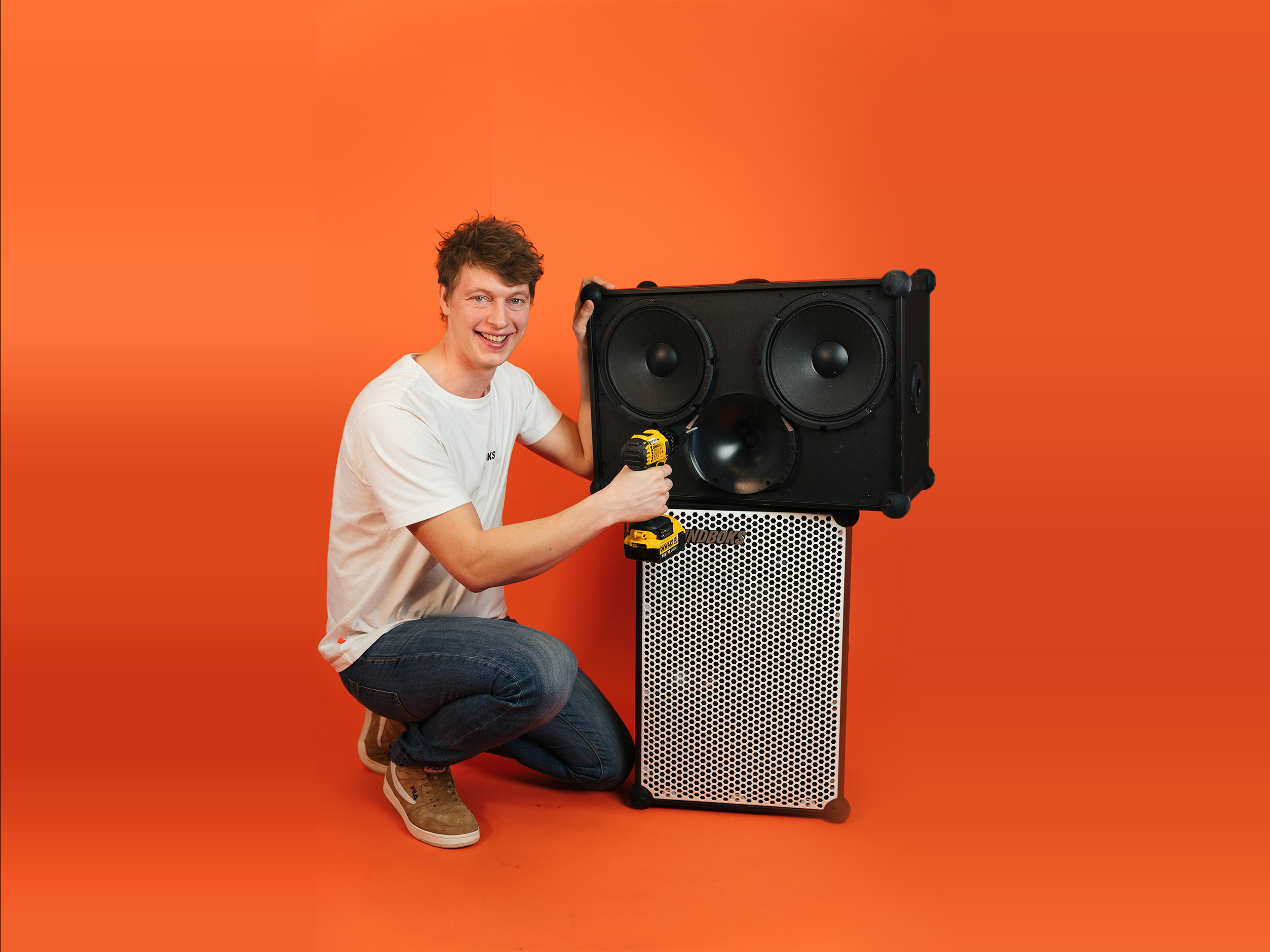 Emil fixing a SOUNDBOKS with a screw driver in front of a SOUNDBOKS in front of an orange background