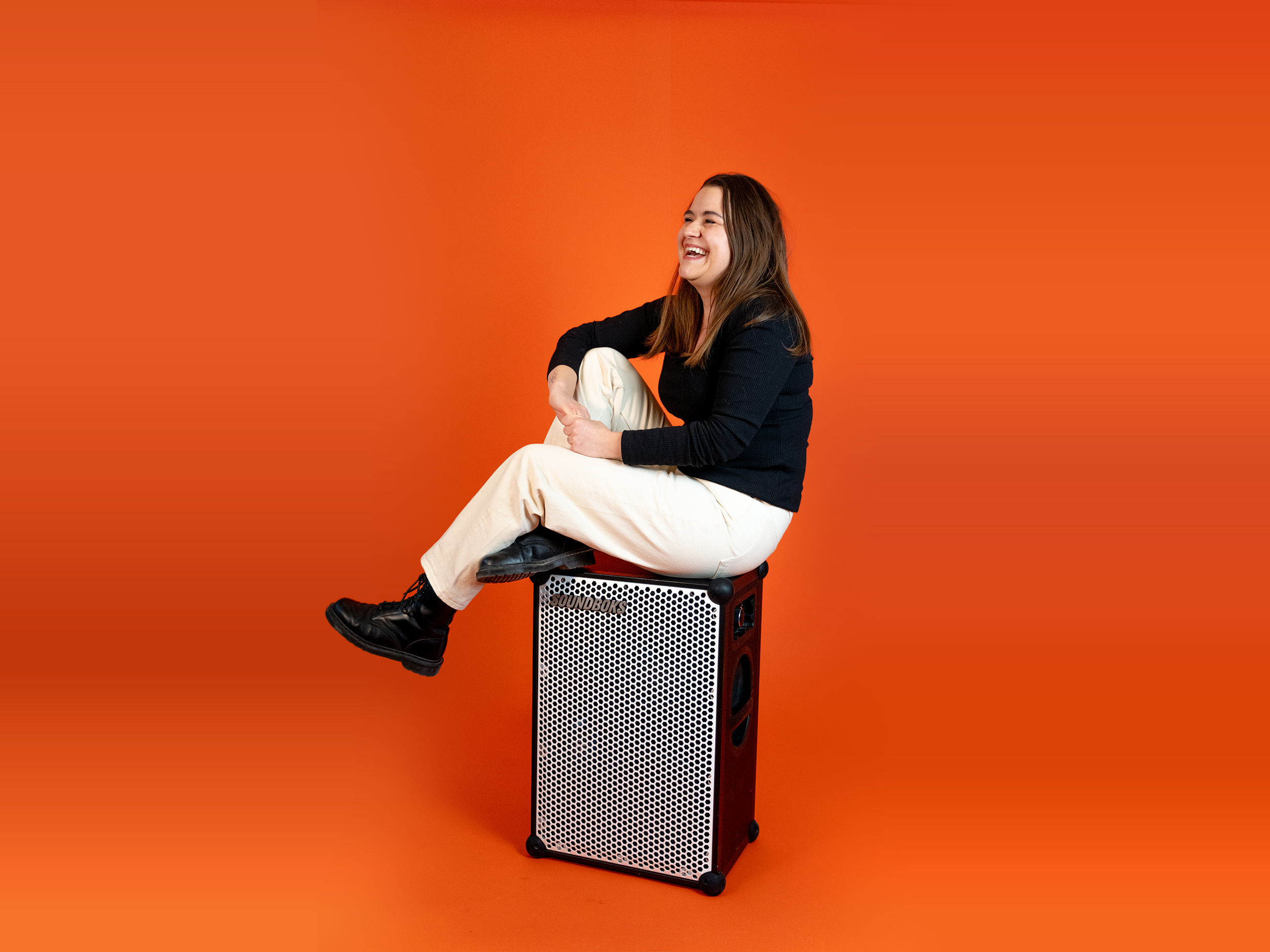 Ewa, in a black outfit sitting on top of a SOUNDBOKS in front of an orange background