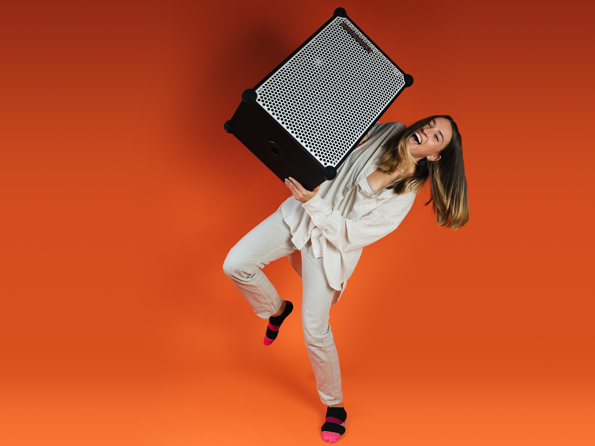 Kait, Brand Experience Team Lead at SOUNDBOKS in front of an orange background with a white SOUNDBOKS in her hands