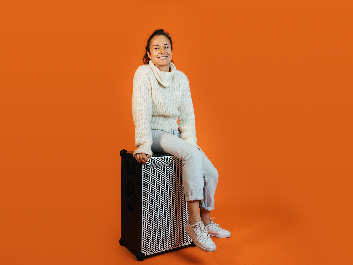 Anjuli, in a white outfit sitting on top of a SOUNDBOKS in front of an orange background