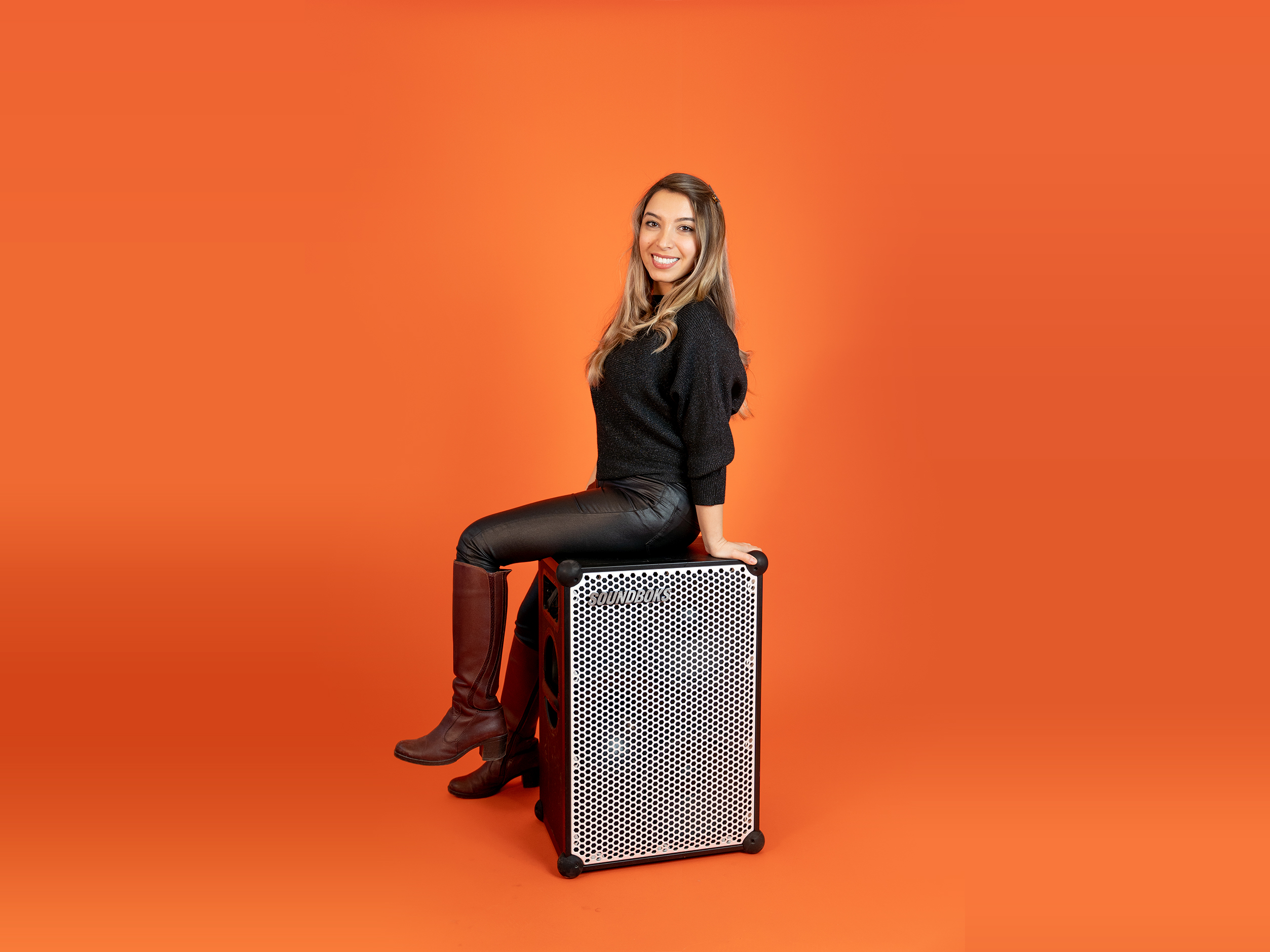 Mehrzad, in a black outfit sitting on top of a SOUNDBOKS in front of an orange background