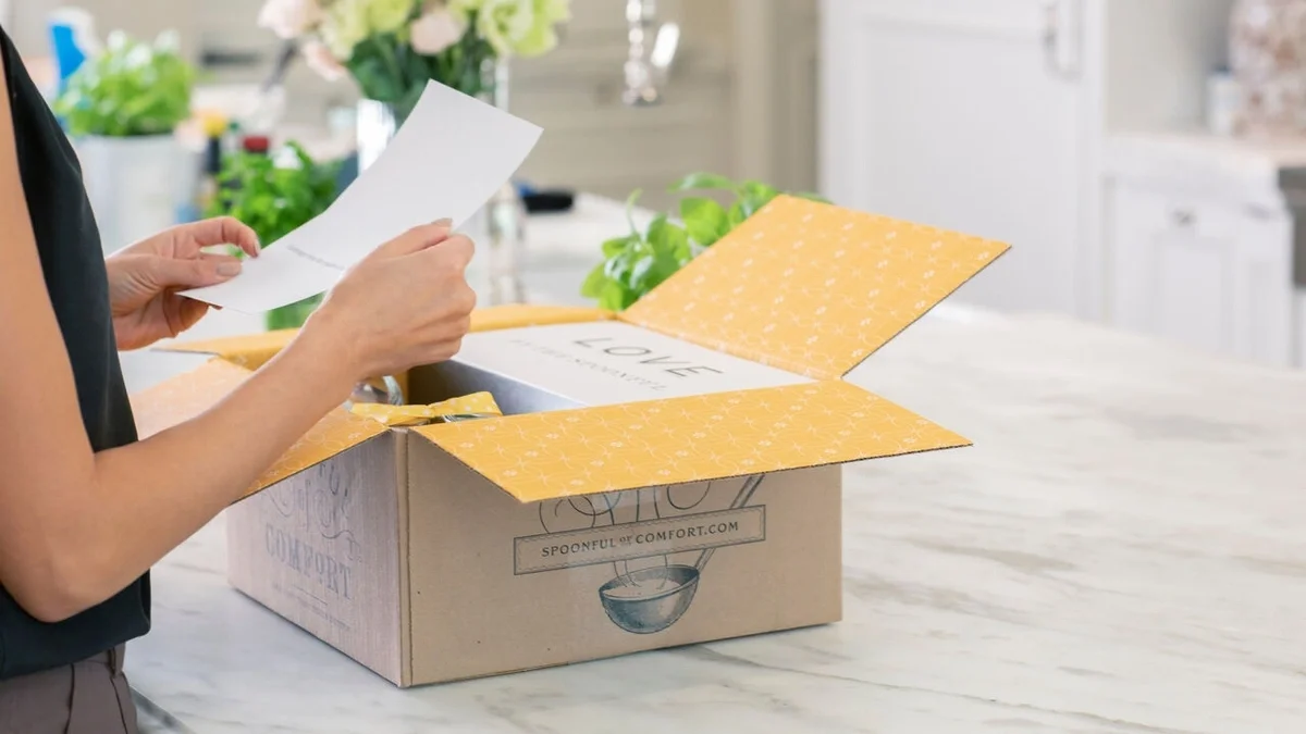 Person opening a Spoonful of Comfort gift box while reading a greeting card in a bright kitchen.