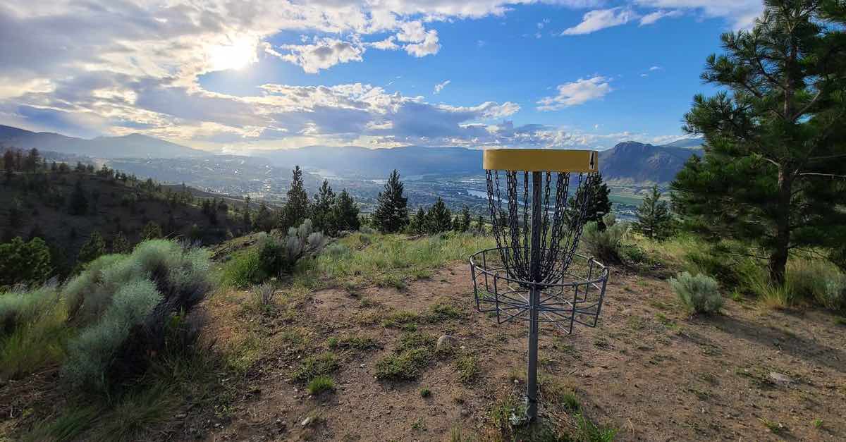 A disc golf basket with a blue sky, sun, and spectacular view of a valley and mountains below