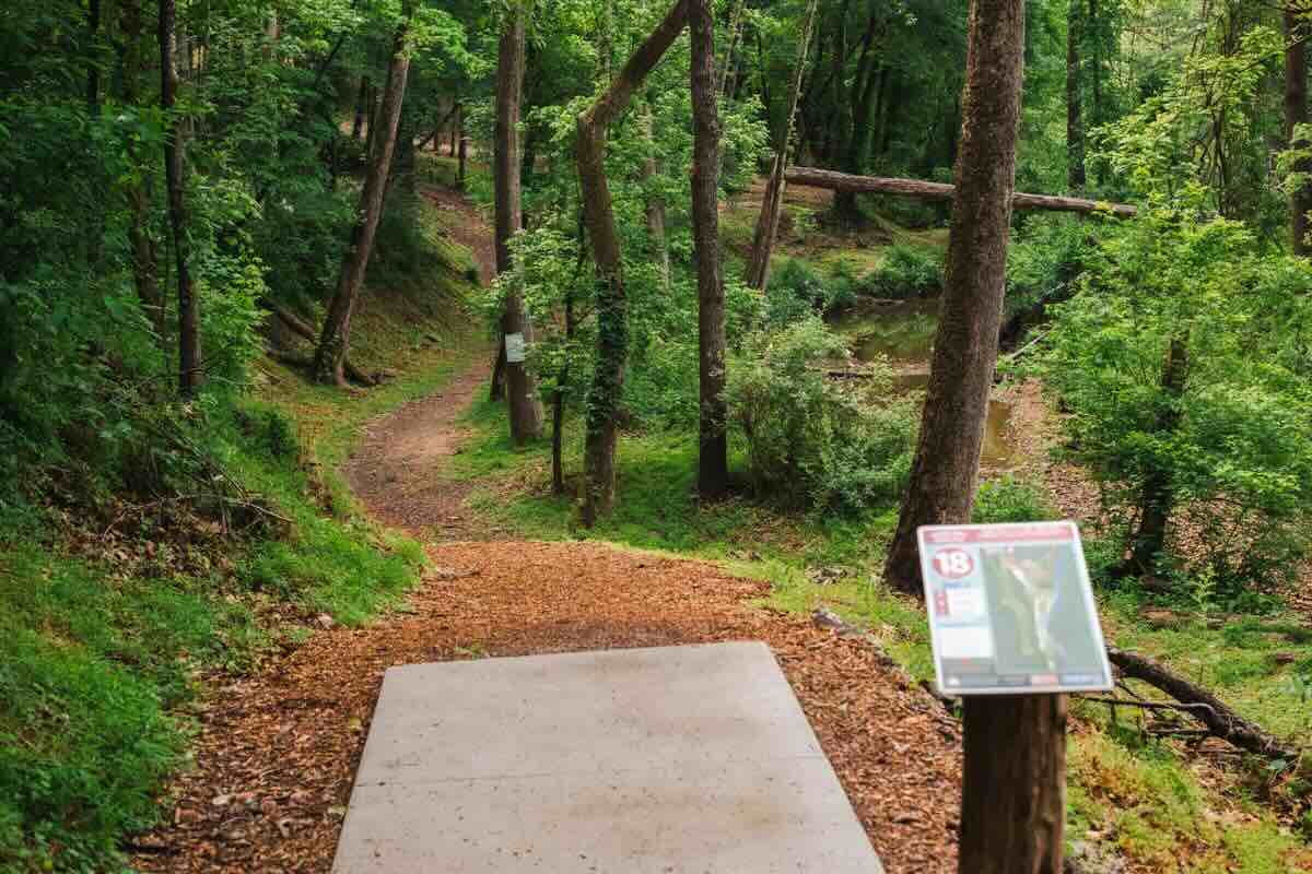 A concrete disc golf tee pad leads to a wooded, curving fairway