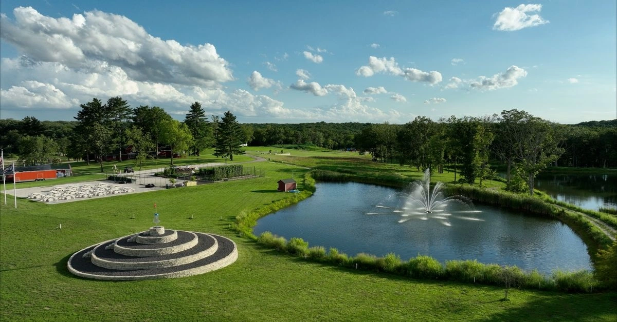 Aerial view of an area with a pond and fountain, multi-tiered construction with a disc golf baskt on top, and mixed mown areas and woods