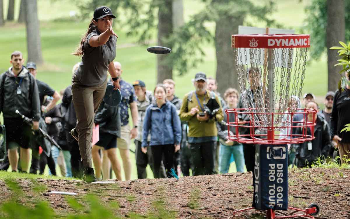 A red disc golf basket with a wrap around the bottom pole woth DGPT and UDisc logos. A woman putts in front of a gallery