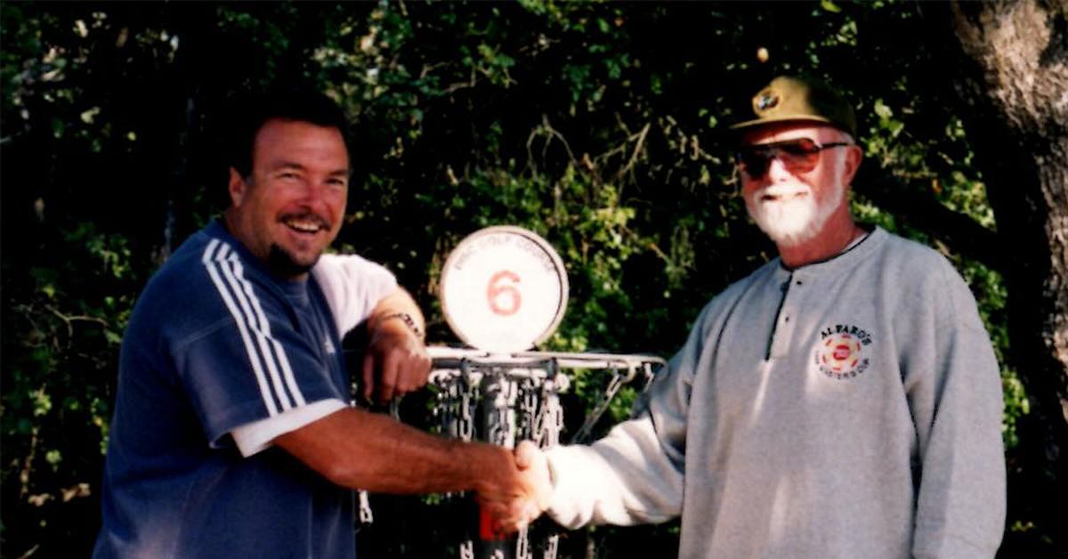 Two men pose for a photo shaking hands in front of a disc golf basket