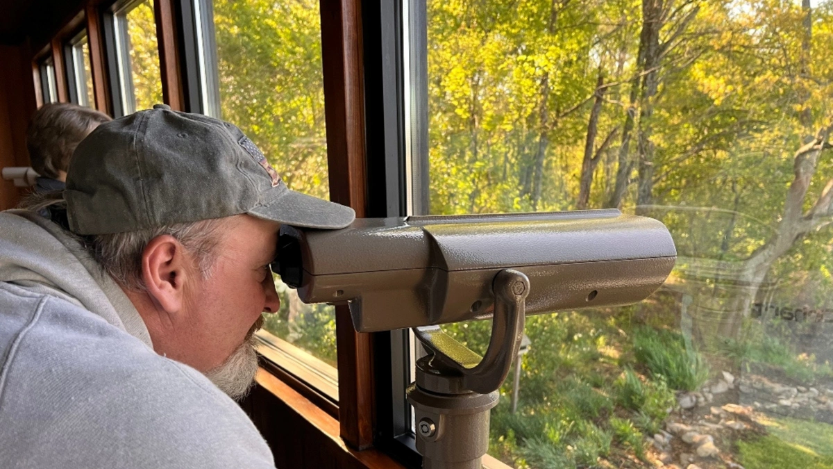 A man in a gray cap looks through a binocular viewer. He’s gazing out a large window that shows a scenic view of trees.