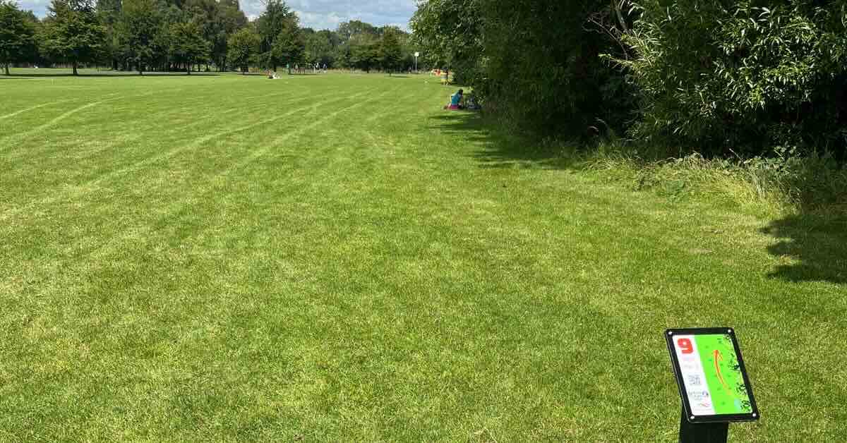 A park of green, mown grass and trees with a disc golf tee sign in the foreground