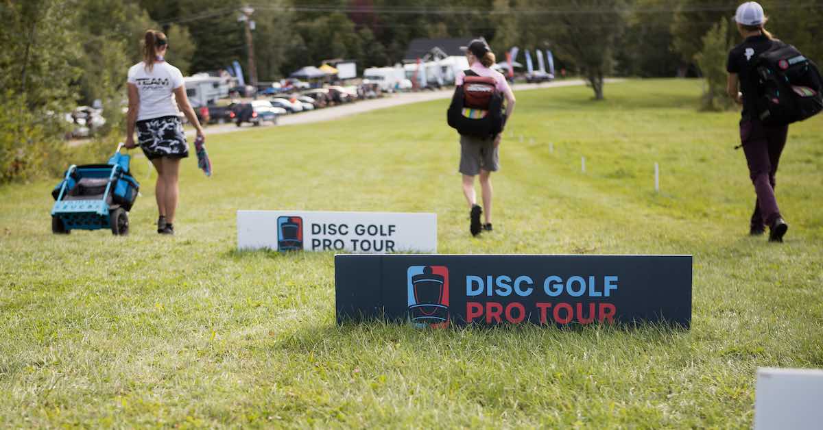 Two signs saying "Disc Golf Pro Tour" on the ground in front of three women walking down a fairway with disc golf equipment