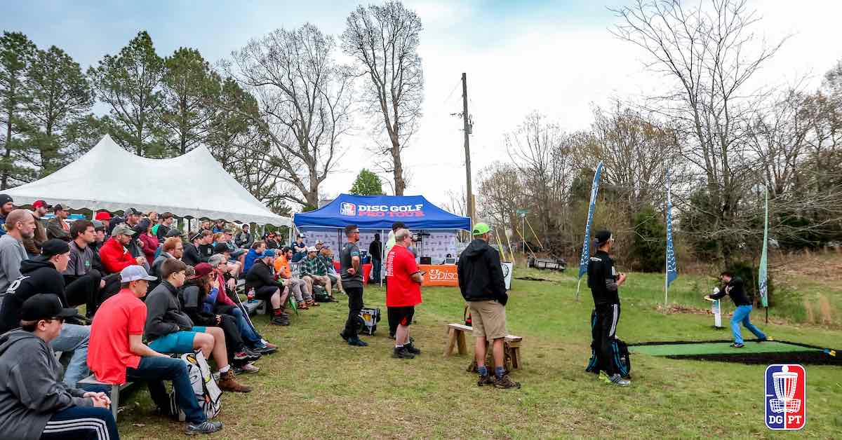 A crowd on bleachers watches someone throw a disc golf drive