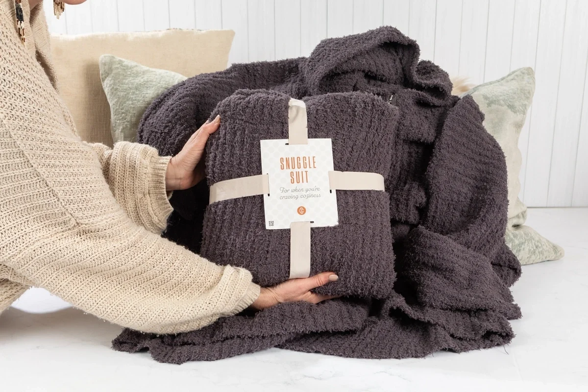 Person holding a folded dark gray plush lounge set with a matching robe and blanket displayed on a soft indoor surface. A tag on the bundle reads “Snuggle Suit.”