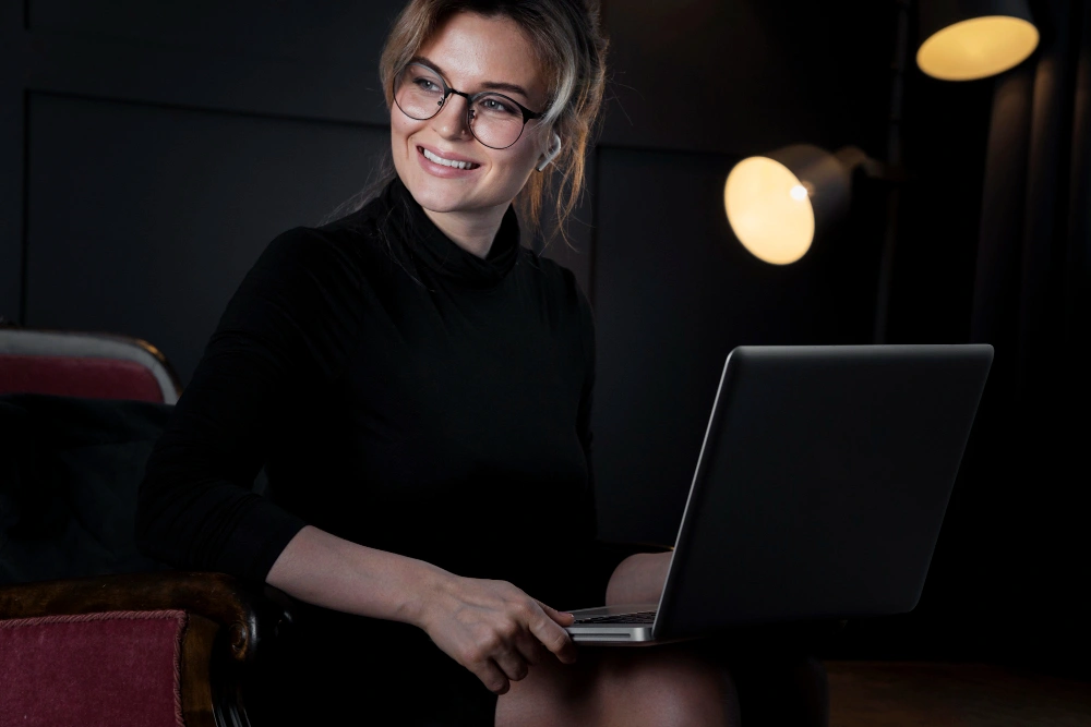 A professional woman sitting with a laptop, smiling, illuminated by soft lighting, representing a confident user working in a tech-friendly environment.