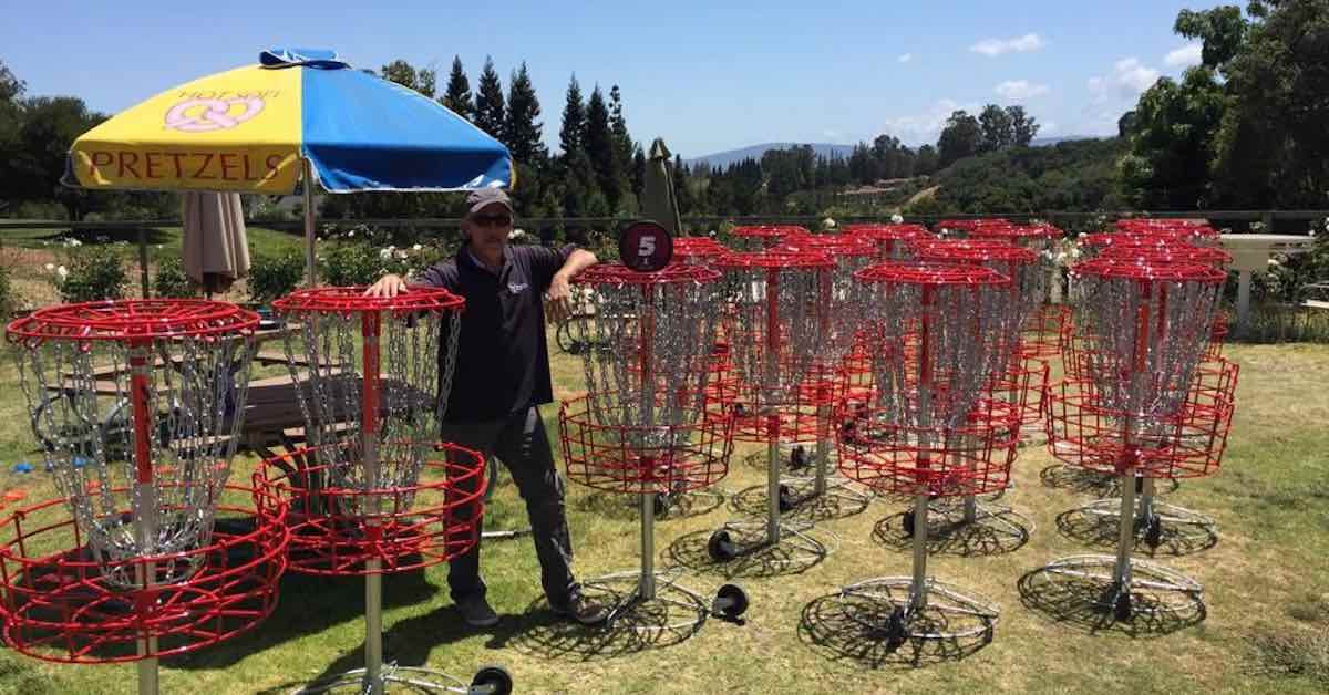 A man stands among many red, mobile disc golf baskets