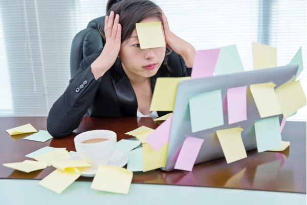 An overwhelmed office worker with hands on her head as she looks at her computer, there are sticky notes everywhere around her.