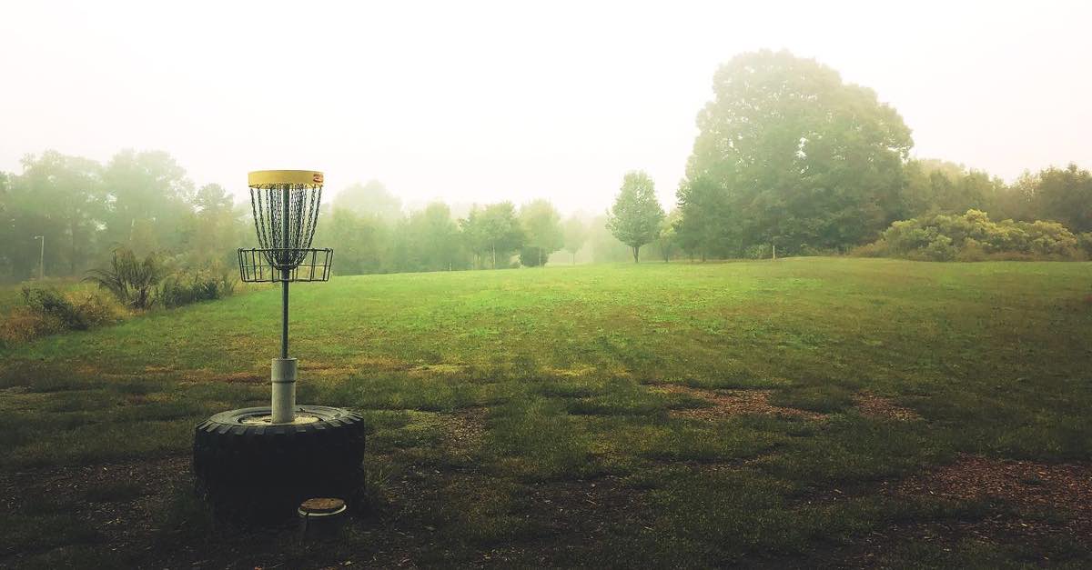 A disc golf basket mounted on large tires in a field on a foggy day
