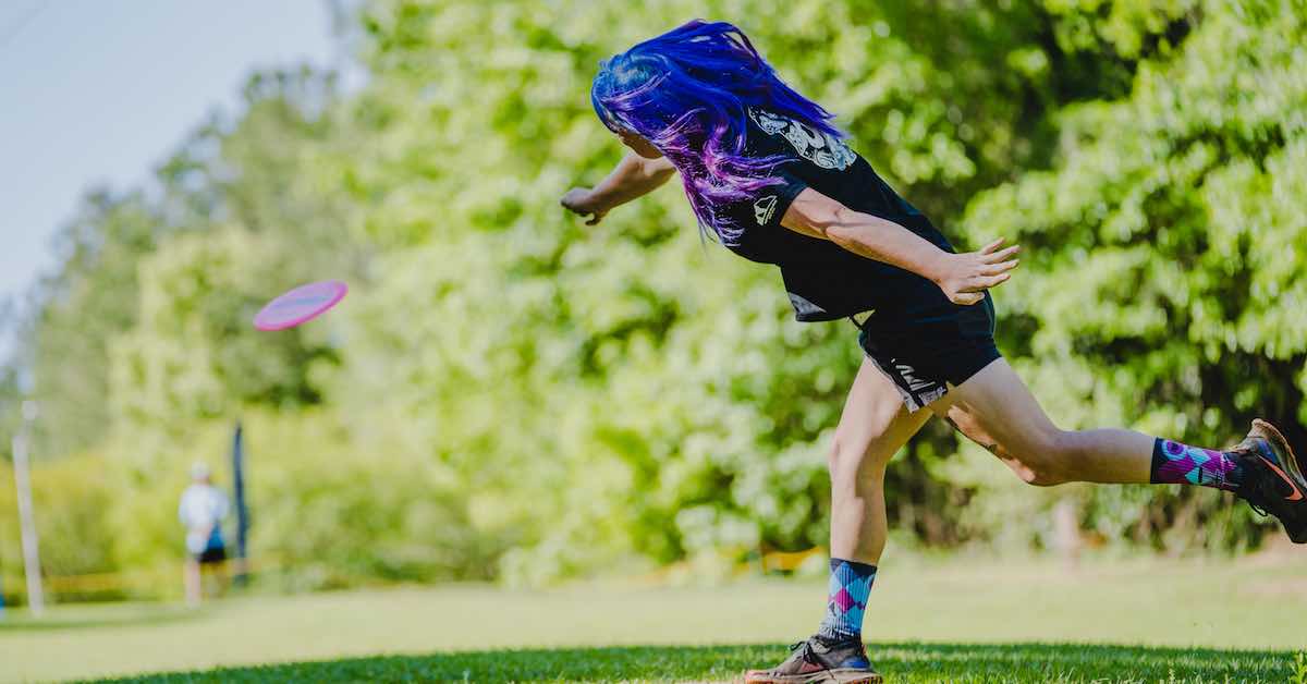 Player with long purple and blue hair after releasing a disc golf drive (side-rear view)