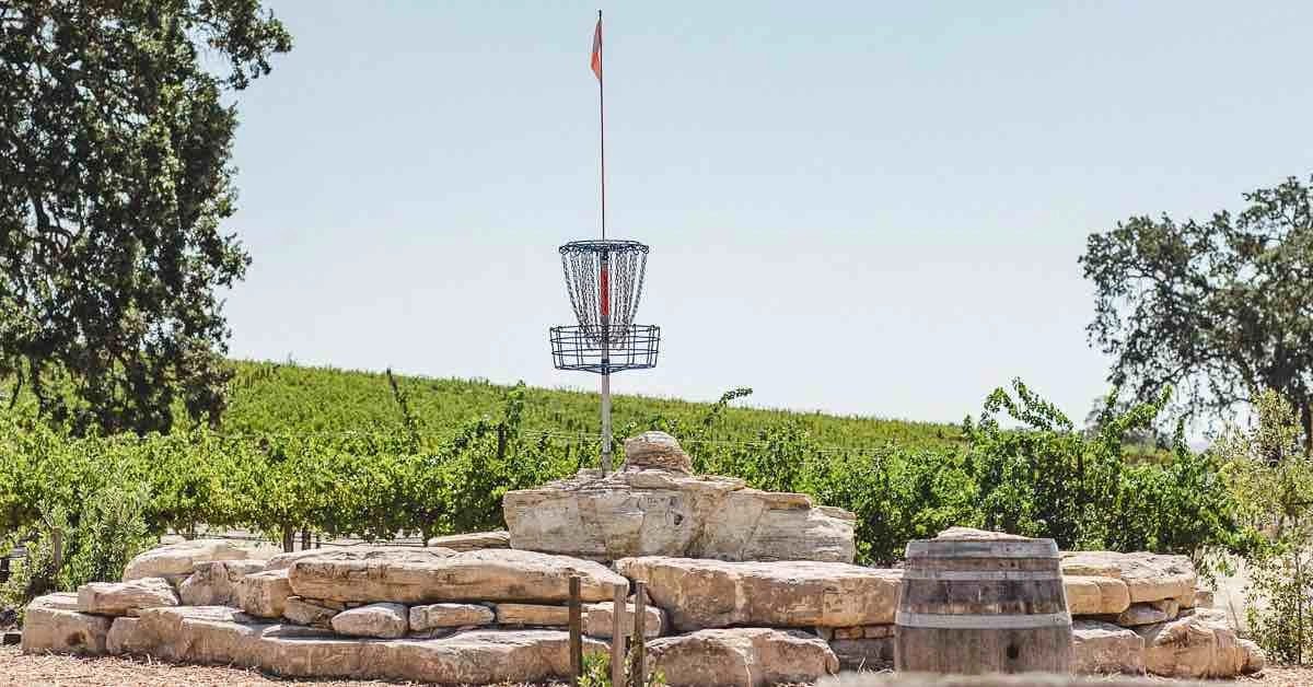 Blue disc golf basket on a rock tower with wine barrels and grape vines in view