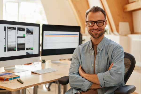 An office worker wearing glasses smiling at the camera while sitting in front of two computer screens happy that he uses a password manager.