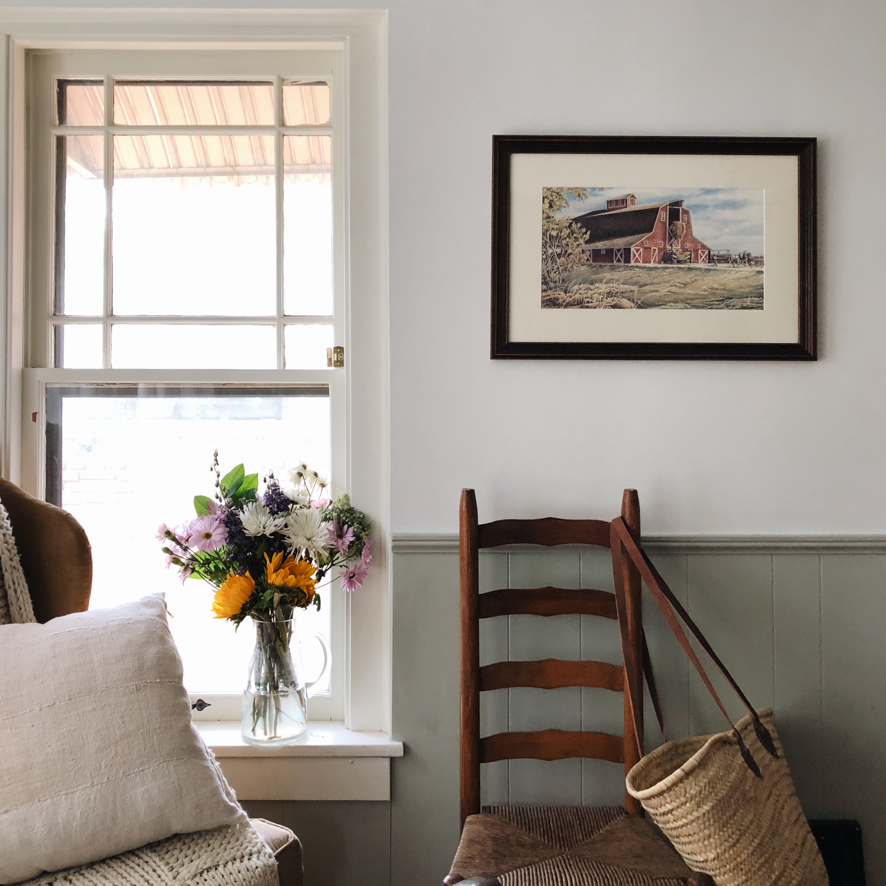 A corner shot of a room with a wicker chair and flowers on the windowsill. A framed drawing of a red barn hangs on the wall in a gold frame.