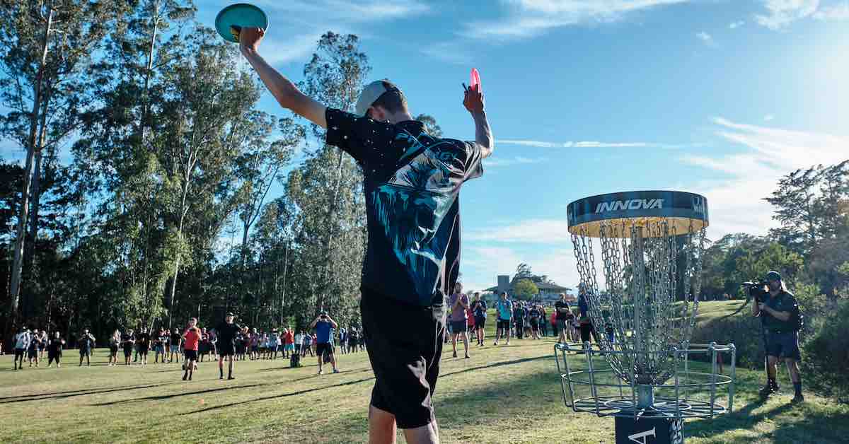 A tall young man holds up his hands in triumph near a disc golf basket in front of a crowd