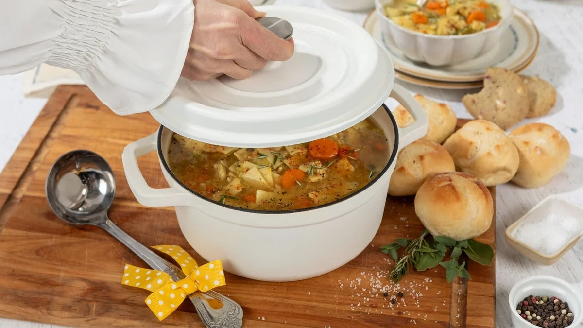 Person lifting the lid from a white Dutch oven filled with chicken soup, surrounded by fresh rolls, a bowl of soup, herbs, and a metal ladle tied with a yellow ribbon.