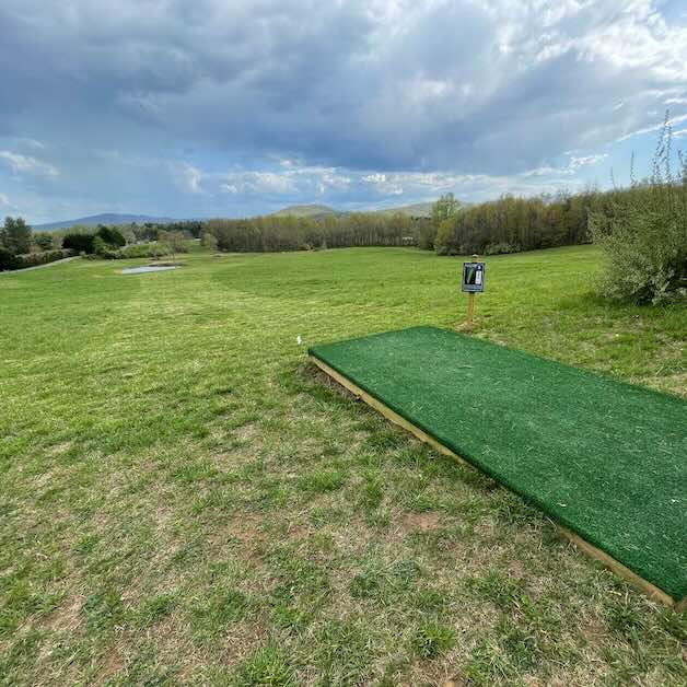 A turf disc golf tee pad leads to a view of well-mown field on a cloudy day