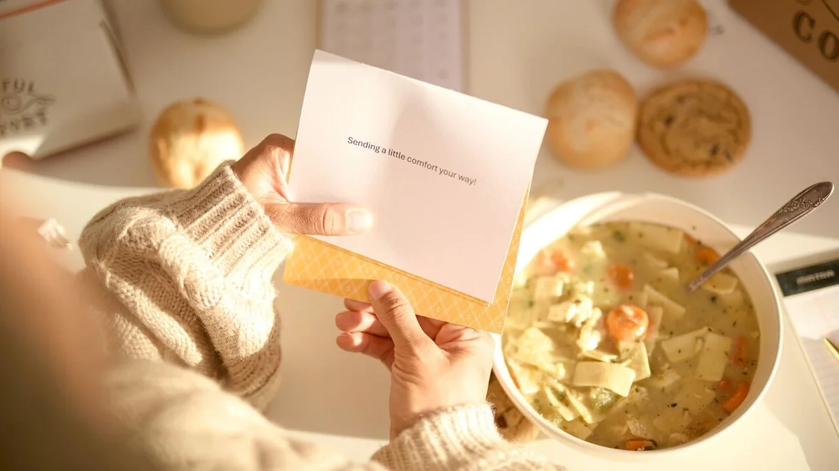 Overhead view of a person in a cozy sweater reading a greeting card while enjoying a bowl of chicken soup, with rolls and cookies nearby in warm sunlight.