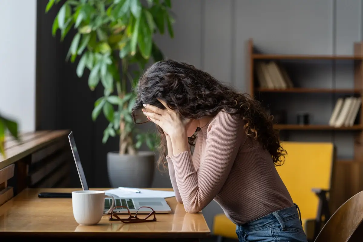 A seated woman with long, curly brown hair holds her heads in her hands over an open laptop