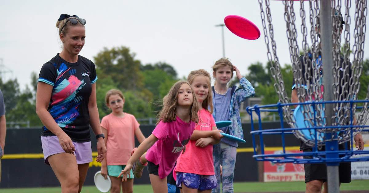 A small girl watching a disc fly toward a basket as others look on