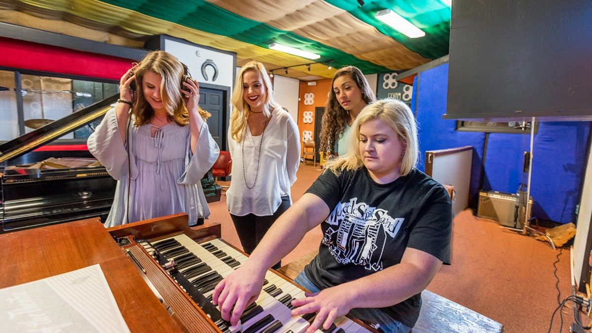 Four women are gathered around a large wooden keyboard. One woman is seated and playing the keyboard, while the other three women are standing and watching.