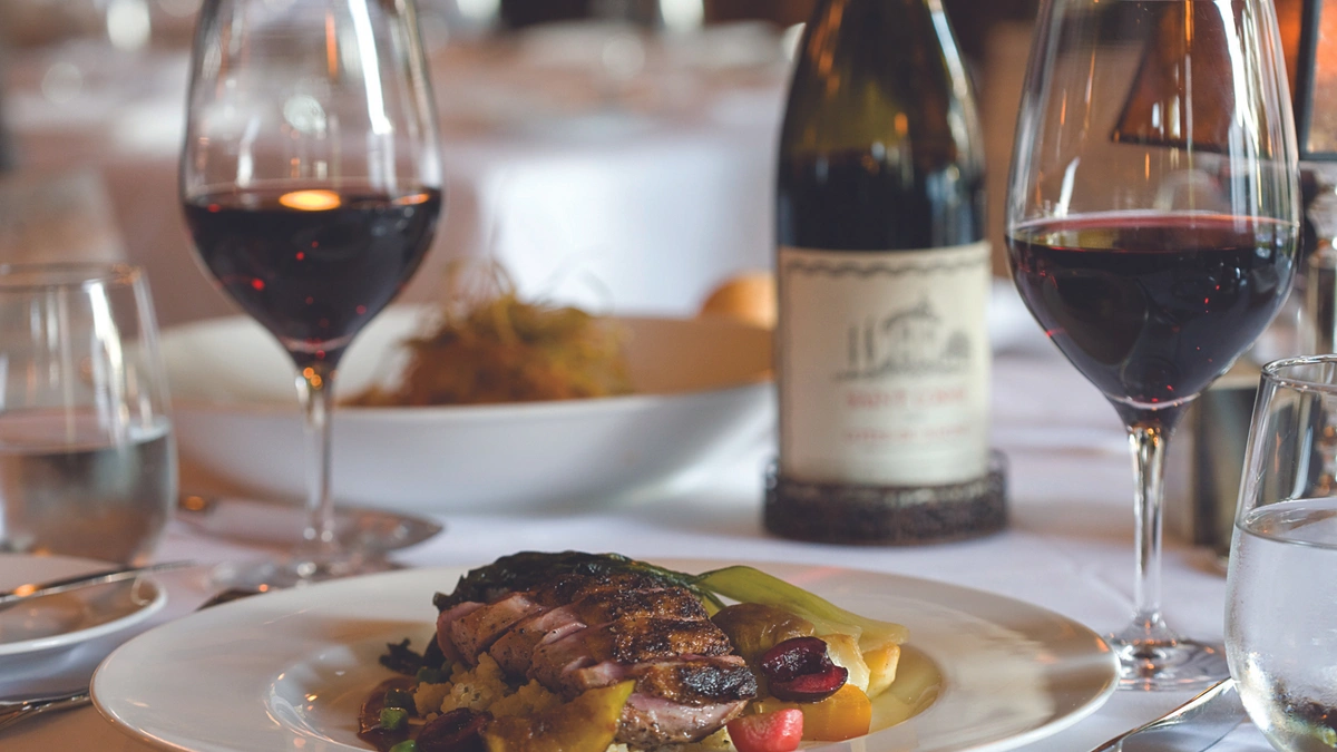 A dining table set with a plate of gourmet food, featuring sliced meat and vegetables, accompanied with red wine.