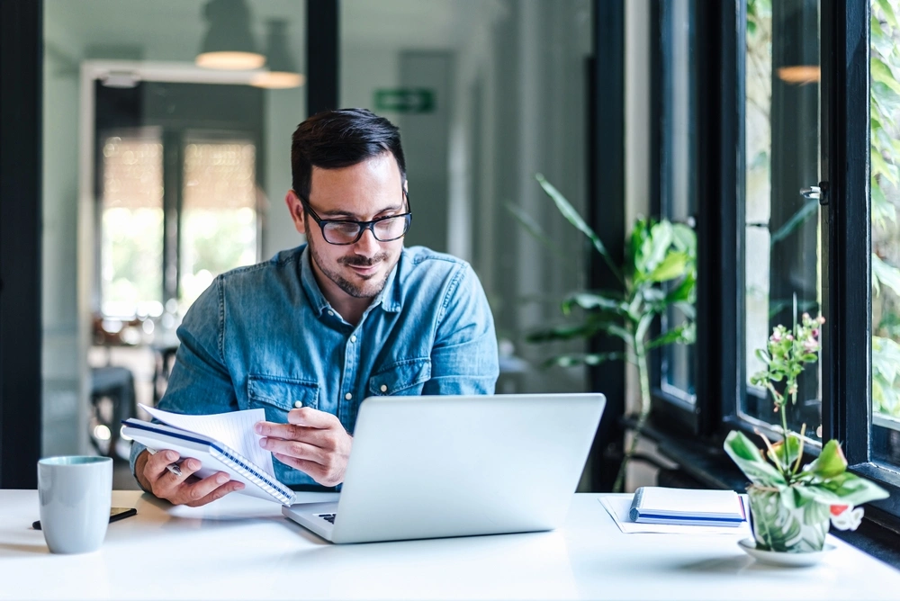 man looking at his computer while holding a notebook