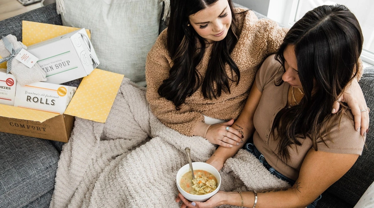 A woman consoles another woman as they both sit on the couch, one eating soups and the other sat next to a Spoonful of Comfort care package