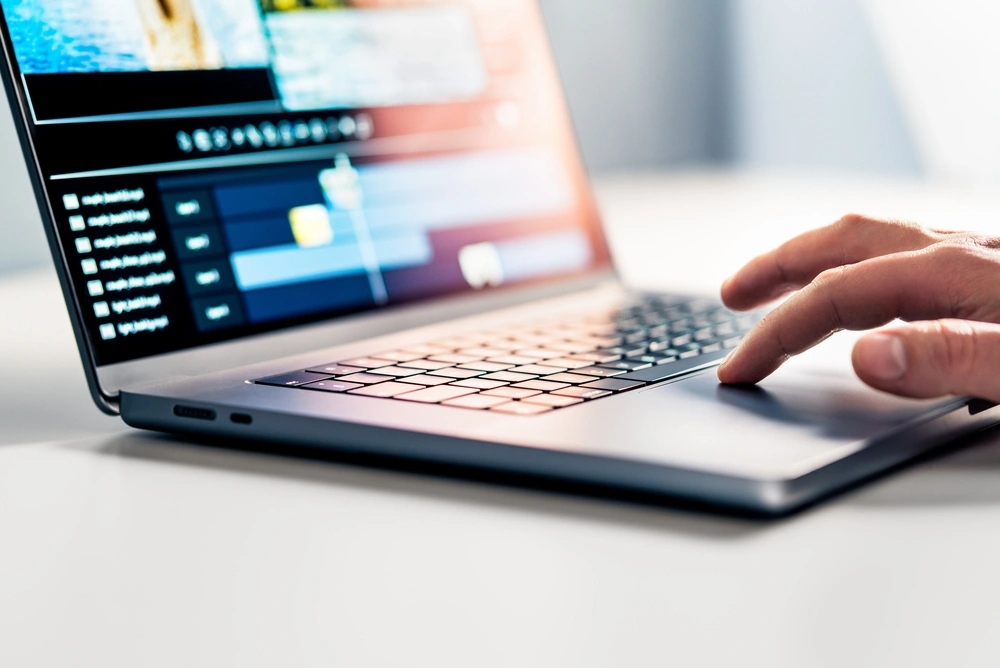 A hand touches the trackpad of a laptop. Editing software is on the screen.
