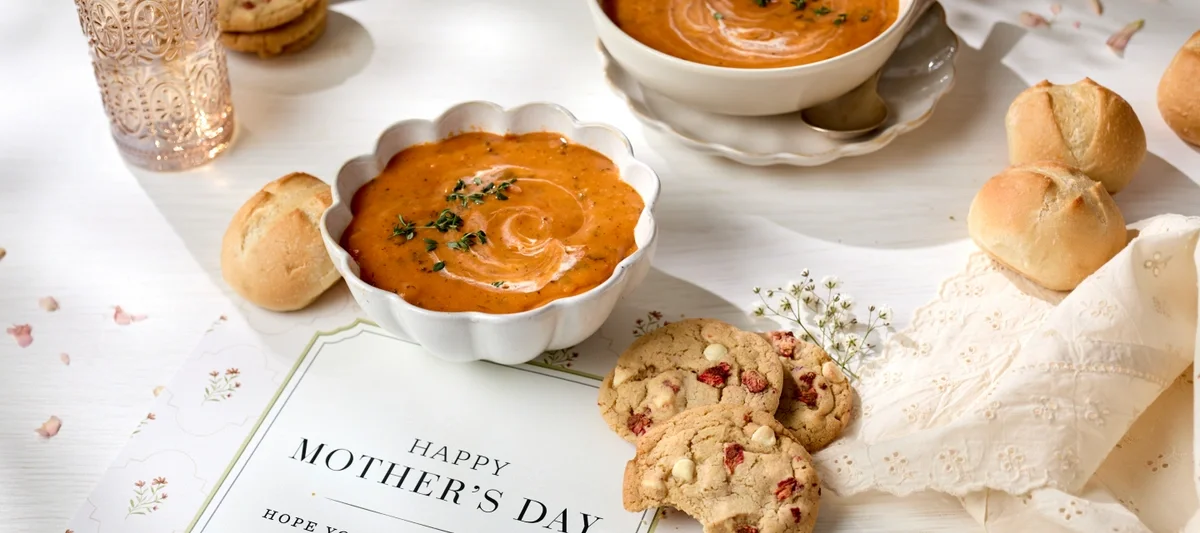 A styled Mother’s Day table featuring two bowls of creamy tomato soup garnished with herbs, fresh bread rolls, and cookies on a white surface, with a card that reads “Happy Mother’s Day — Hope your day is soup-er.”