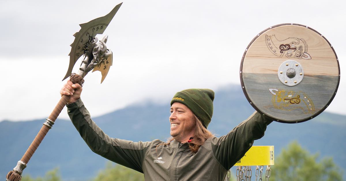 A man dressed for cold weather smiles while holding up a shield and elaborate axe