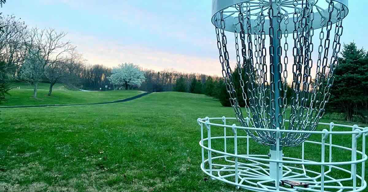 A disc golf basket in a well-mown area with scattered trees at sunrise or sunset