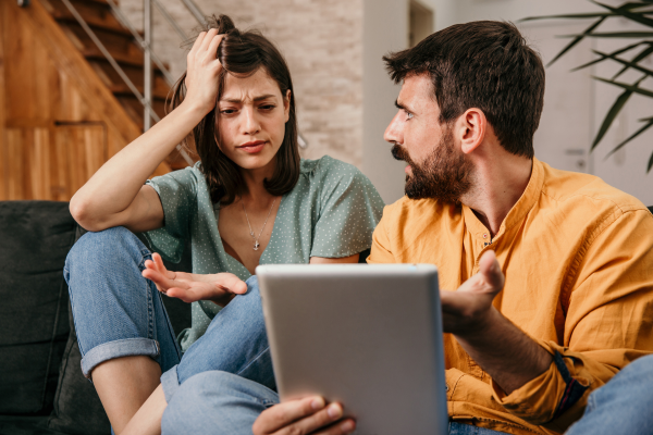 A man and woman are sitting down together. The man is holding a computer and looking at the woman, while the woman holds her head in confusion looking whilst looking at the computer.