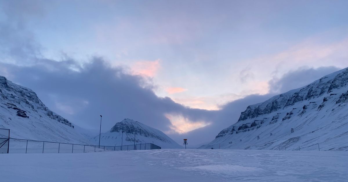 A stark landscape of snow, mountains, and a fence with a lonely disc golf basket