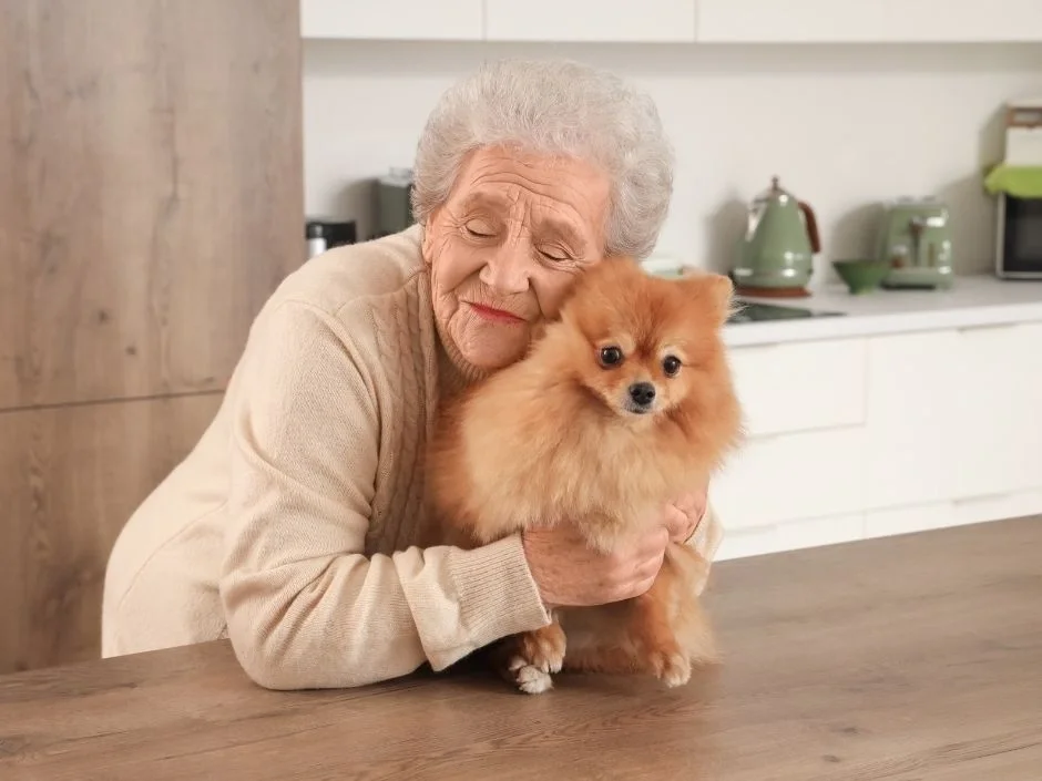 Elderly woman with closed eyes holding a Pomeranian close in a peaceful home kitchen