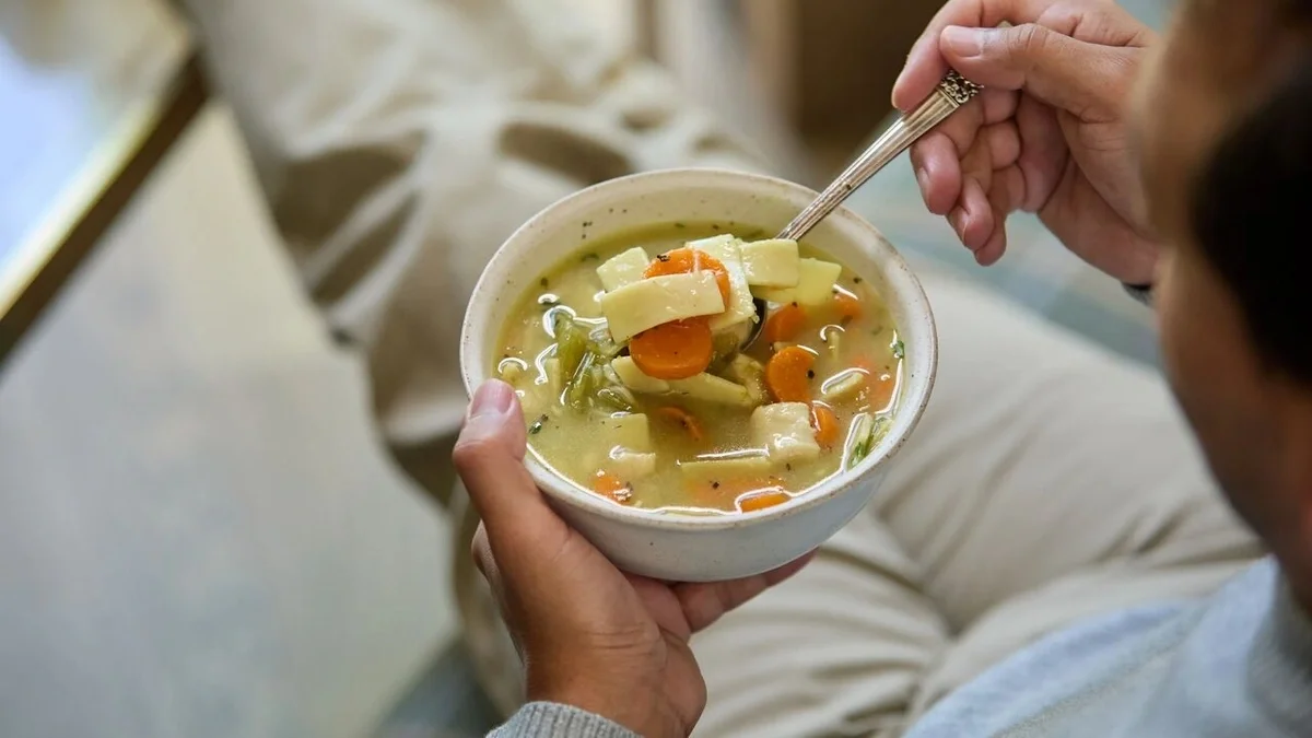 Close-up of a person holding a bowl of chicken soup with carrots, celery, and noodles while lifting a spoonful.