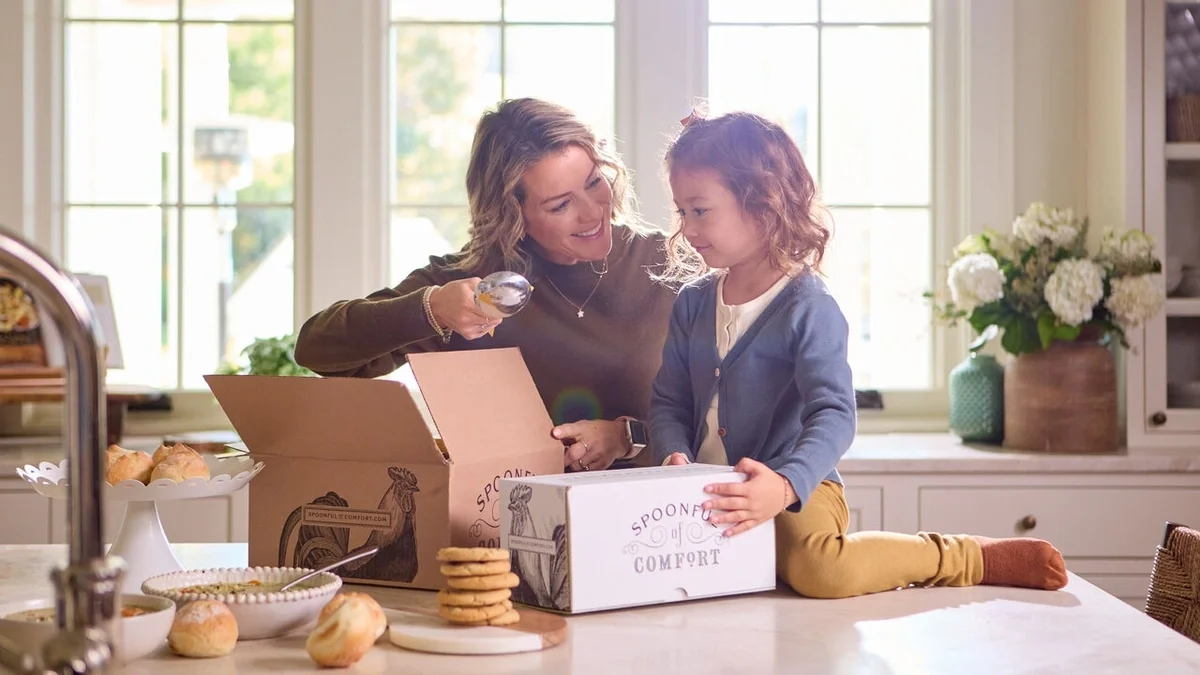 Mother and young child smiling together in a bright kitchen as the mother playfully holds a spoon near the child's mouth. An open Spoonful of Comfort gift box, rolls, cookies, and a bowl of soup sit on the counter.