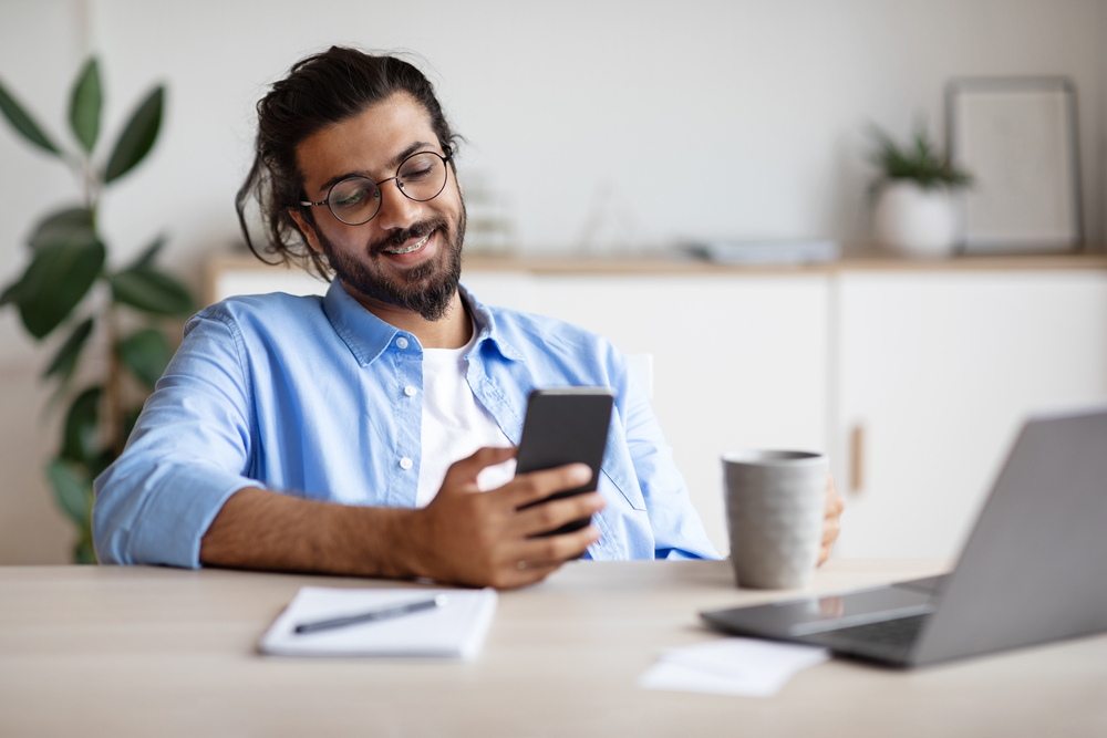 Happy Indian man using his phone and laptop drinking coffee