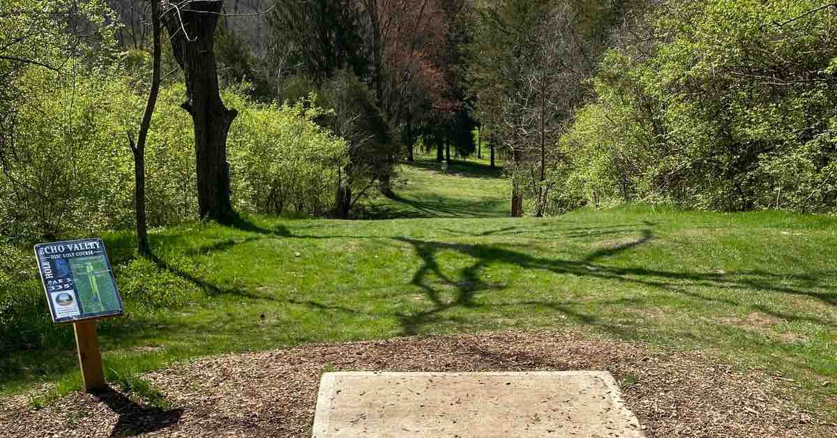 A concrete tee pad leads to a grassy fairwy lined by trees