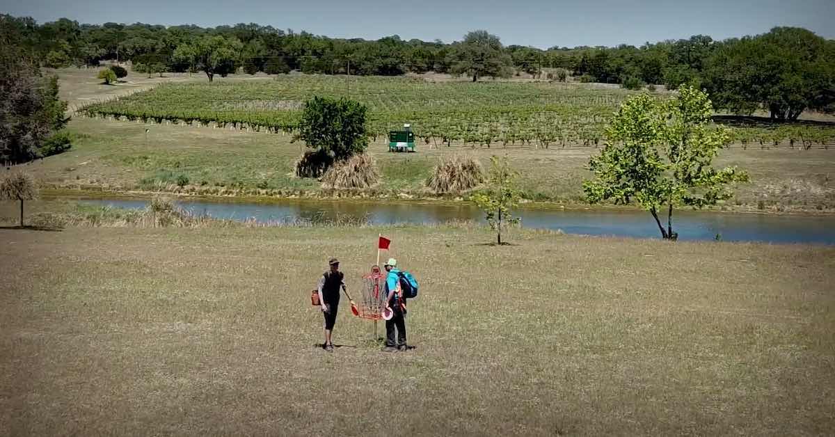 Two disc golfers at a basket and over a small body of water are rows of grapevines