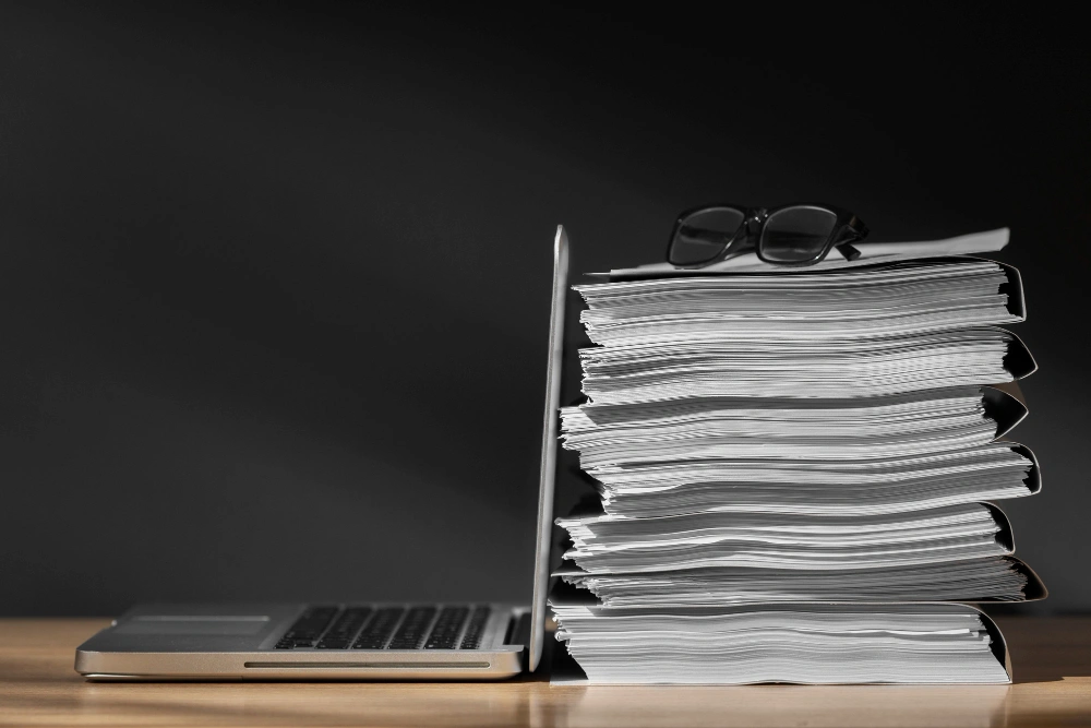 A laptop partially obscured by a large stack of documents with a pair of glasses resting on top.