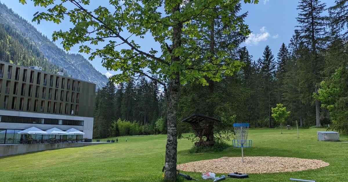 A blue disc golf basket in a mowed area with a hotel to the left and mountains beyond