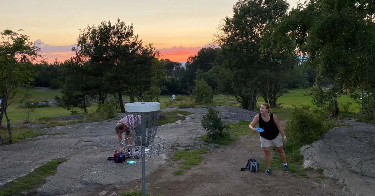 A woman putts at a disc golf basket in an area of grass and exposed rock at sunset