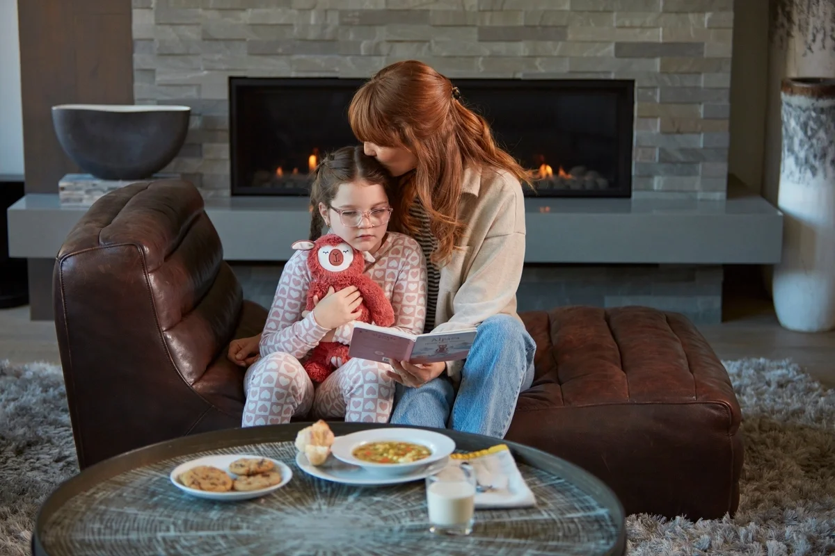 A woman and young girl sit together on a brown leather couch in front of a fireplace, reading a book. The child holds a stuffed animal while a bowl of soup, cookies, and a glass of milk rest on a coffee table in the foreground.